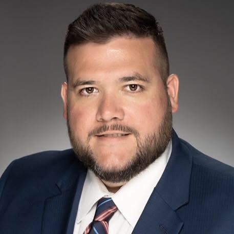 Close up headshot of Chris with dark background wearing a dark blue suit, white collar shirt and tie and his hair is short and clean cut
