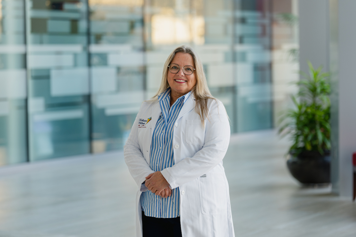 Photo of Debbi standing in bright lobby area with hands cross at waist wearing her blonde hair down, glasses, a white doctors lab coat and blue and white vertical stripe shirt with collar and black pants