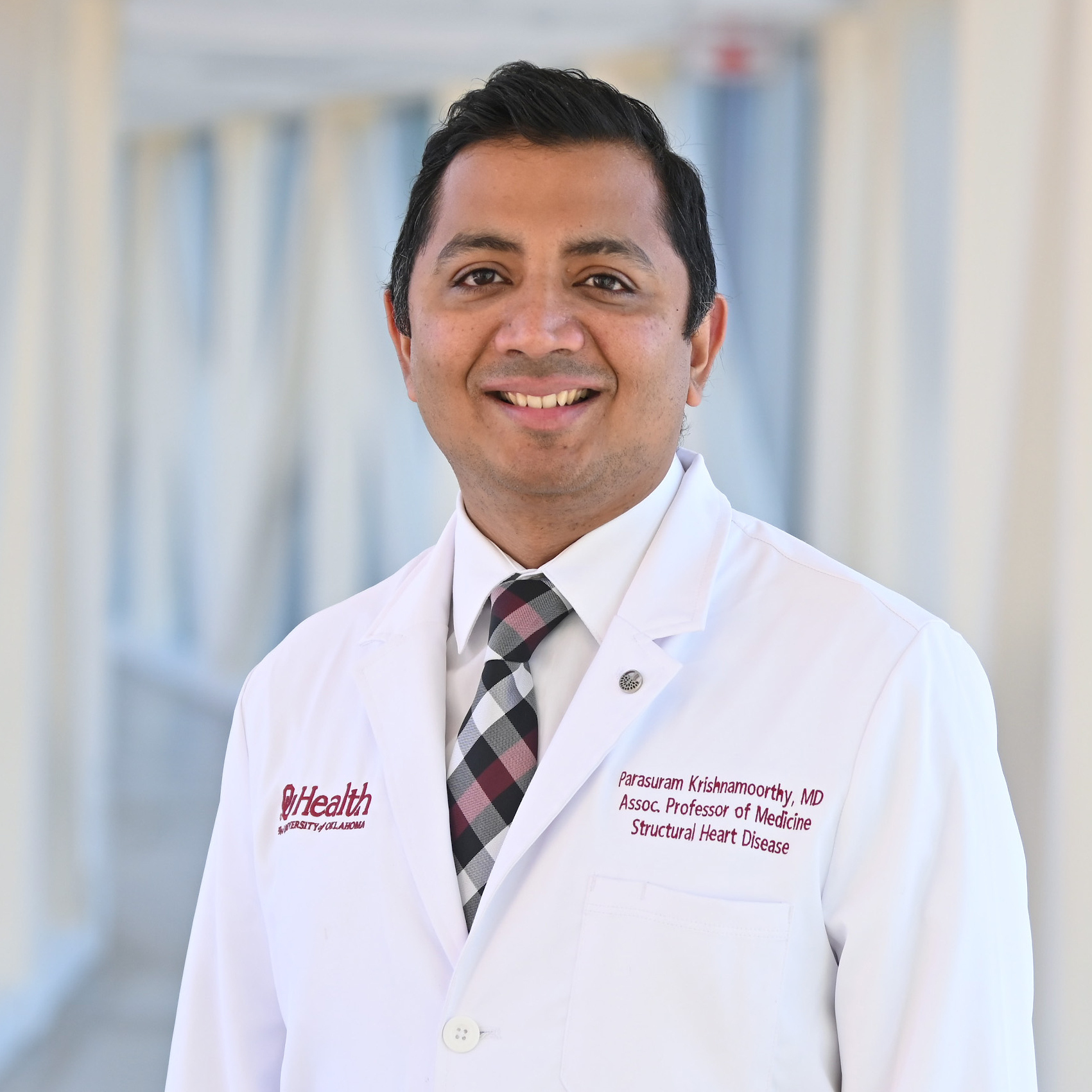 Professional headshot of a healthcare provider wearing a white OU Health lab coat and patterned tie, standing in a bright, softly blurred hospital corridor.
