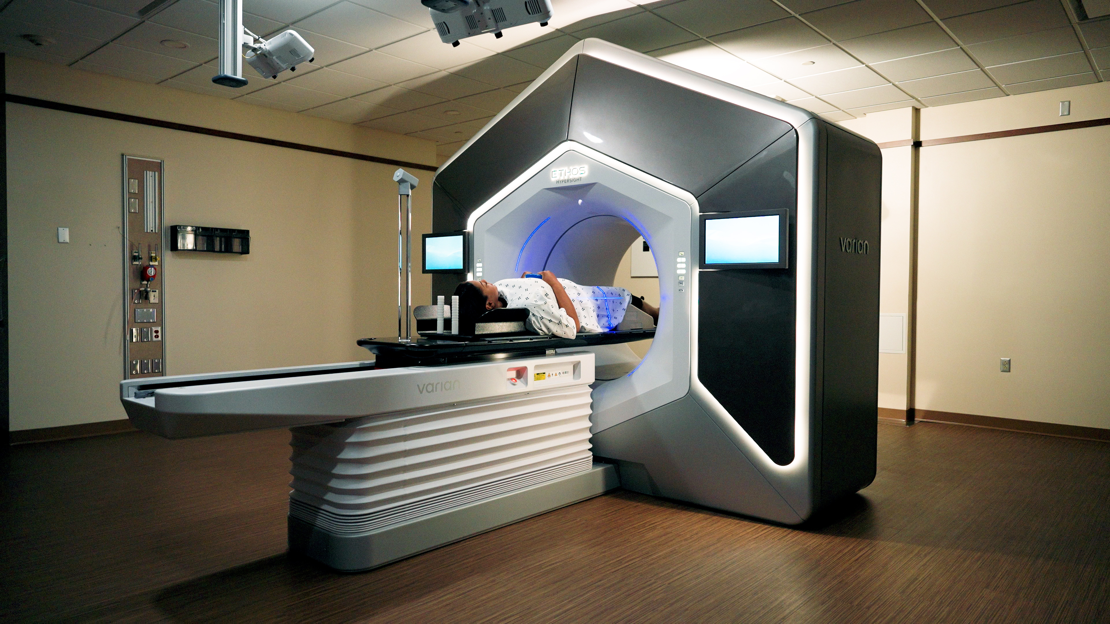 A patient lies on a treatment table positioned within a Varian ETHOS radiation therapy machine, surrounded by illuminated imaging equipment in a hospital treatment room.