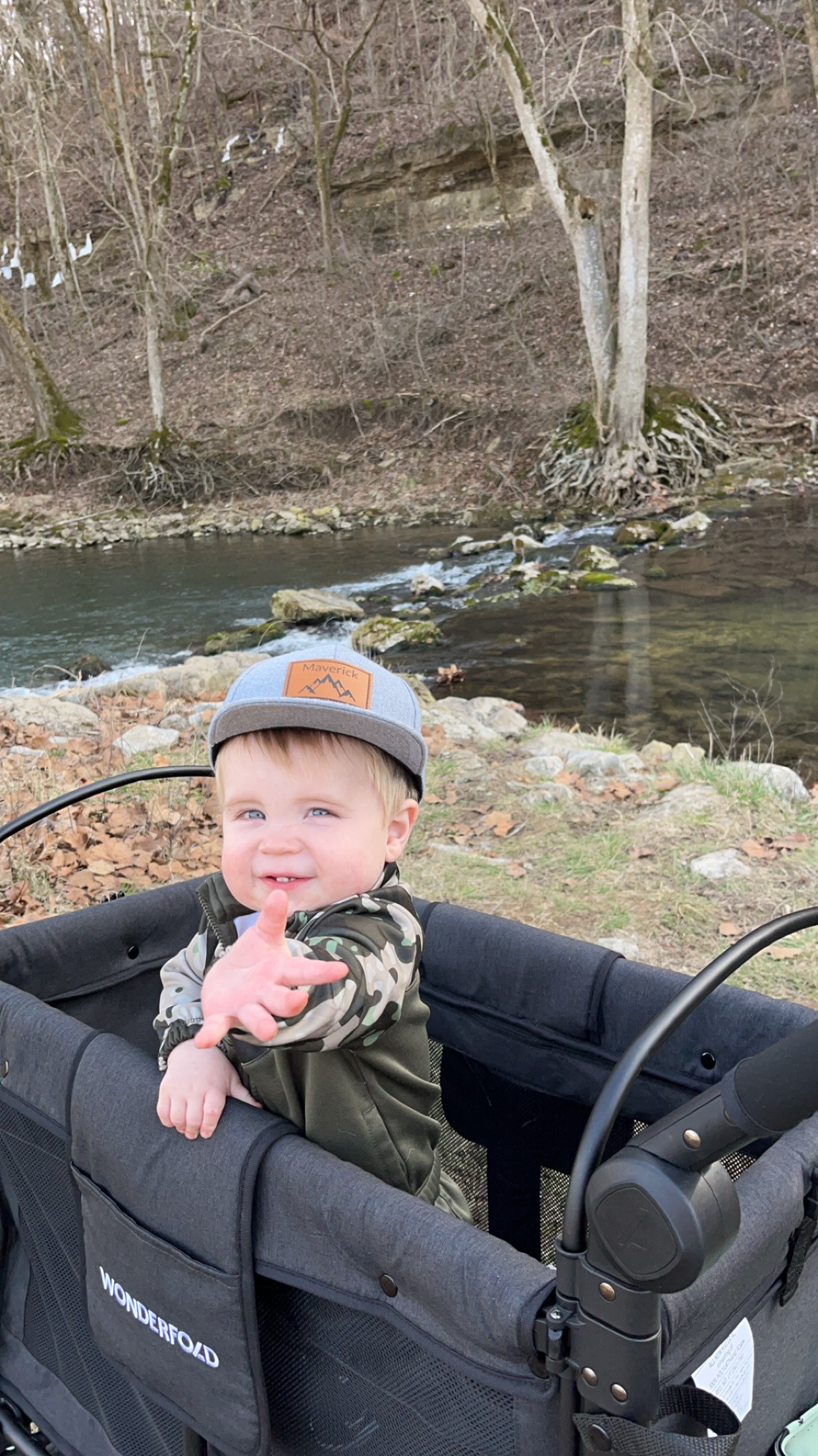 Young child sitting in a stroller wagon beside a shallow creek, reaching toward the camera while outdoors in a wooded area with rocks, trees, and water in the background.