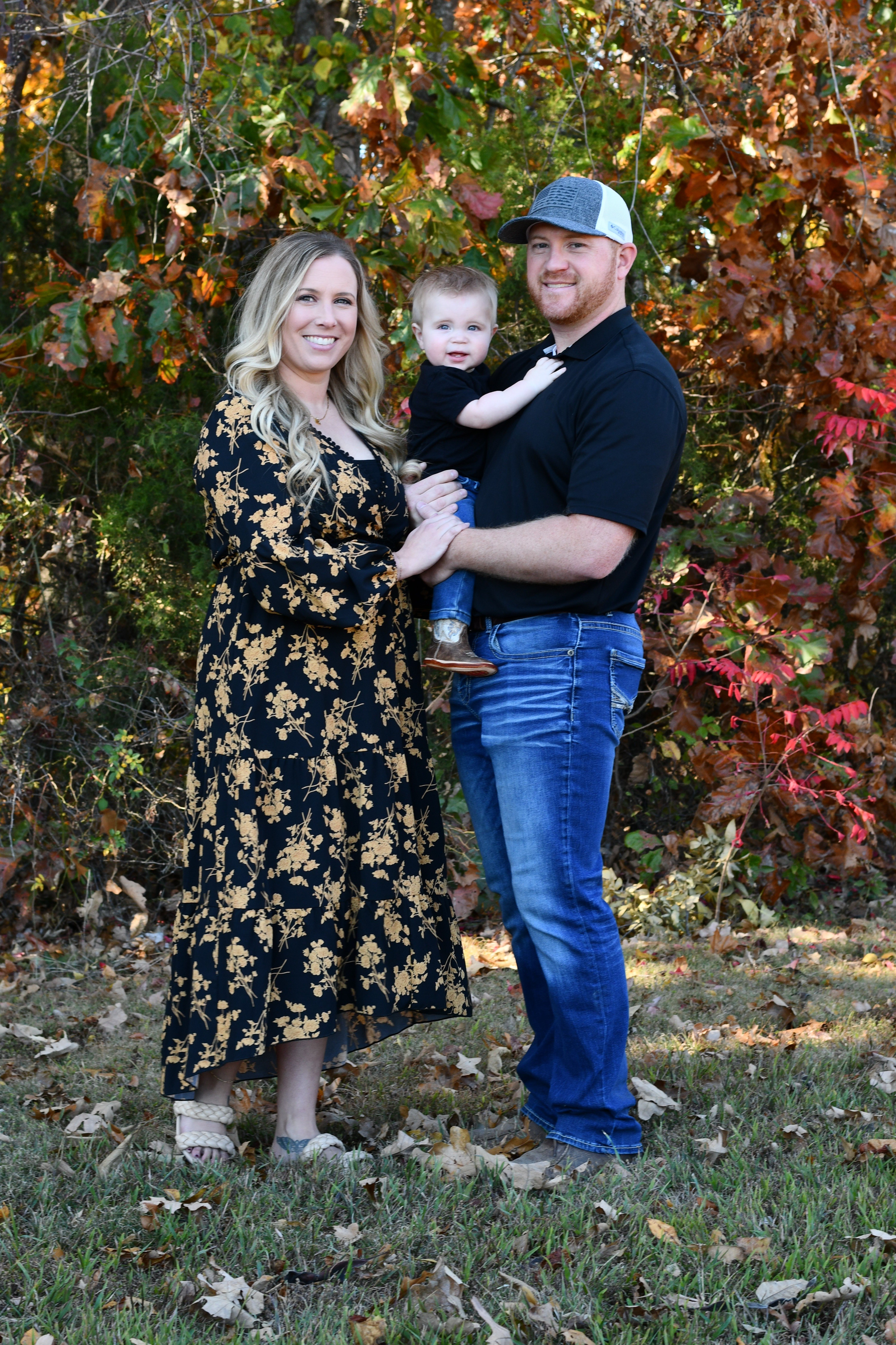 Family of three pose outdoors in front of colorful autumn foliage, with one adult holding a young child while another adult stands beside them. The group faces the camera in a family portrait setting.