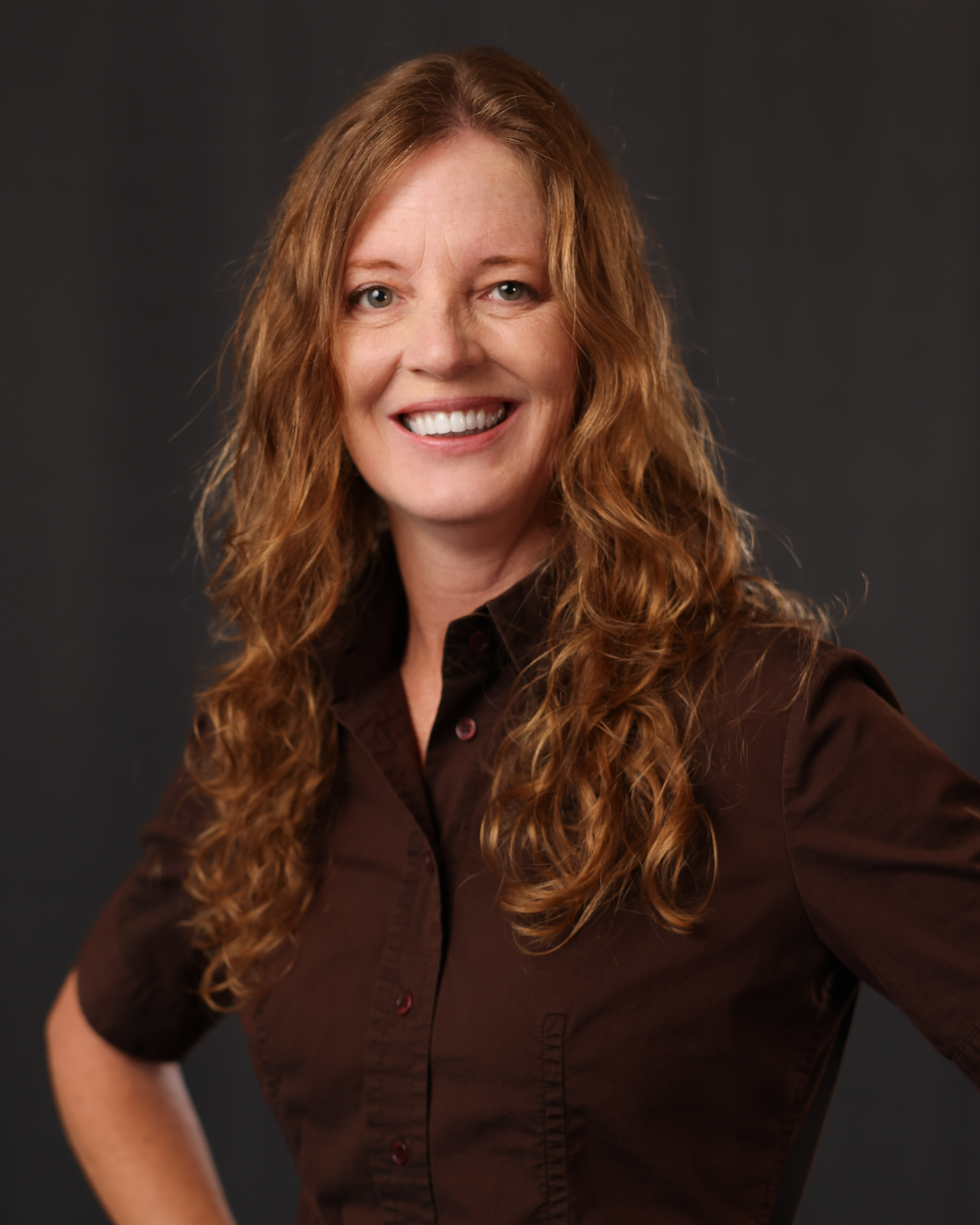 Headshot of Megan with dark background with her red hair down and over both her shoulders wearing a dark brown button up collar three quarter shirt with her hands on her hips 