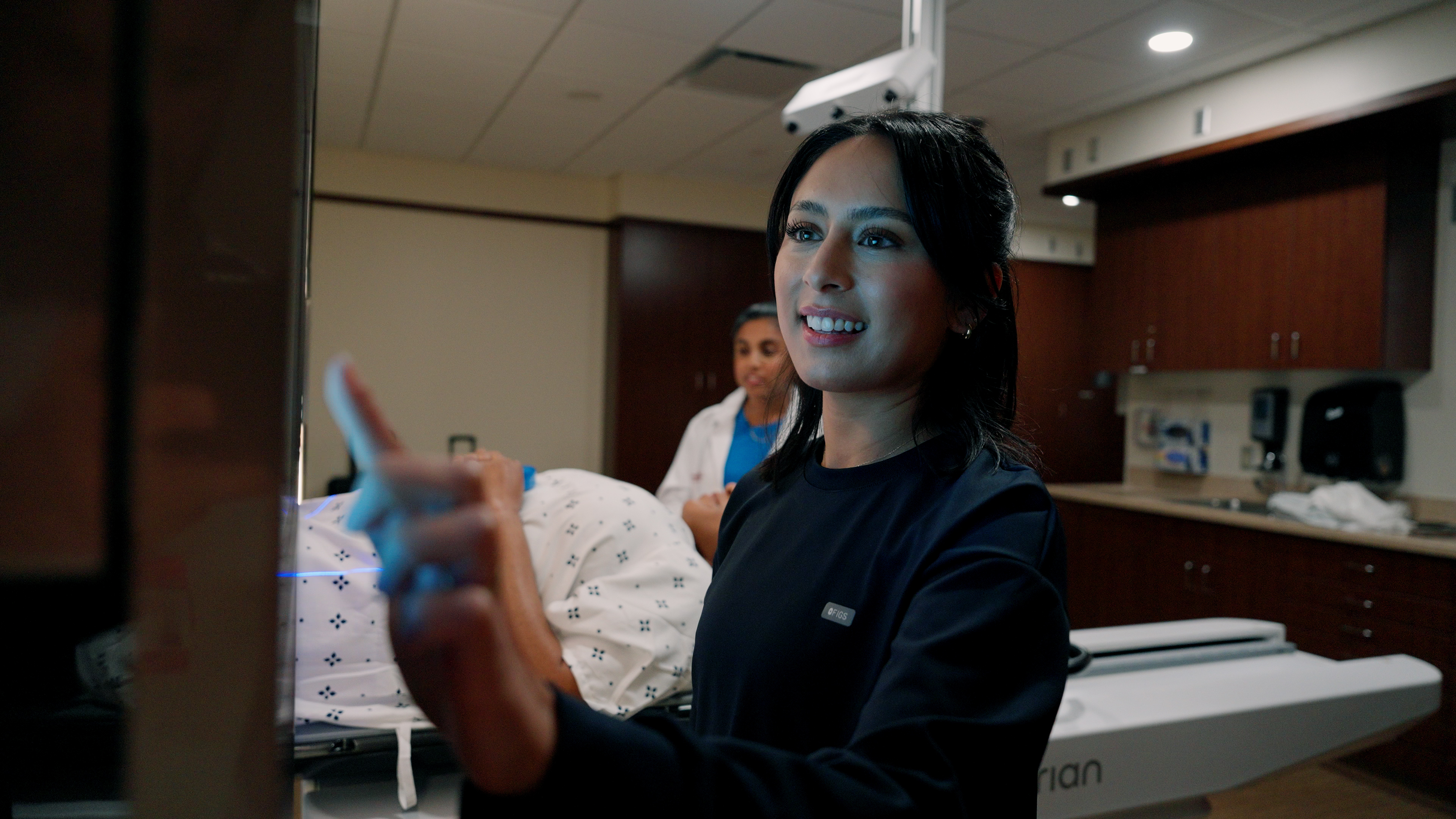 A healthcare professional operates imaging or treatment equipment beside a patient lying on a treatment table, with another clinical staff member visible in the background of a modern treatment room.