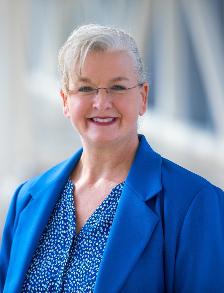 Close up headshot of Tracy with neutral background with her hair styled up wearing a turquoise blazer and a matching shirt with a speckled pattern