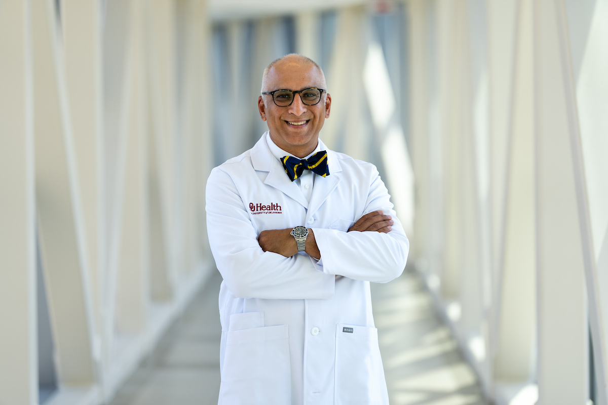 Headshot of Dr. Ahmed standing in front of neutral background with arms crossed while smiling and wearing glasses, bow tie, white doctors lab coat and watch on left wrist