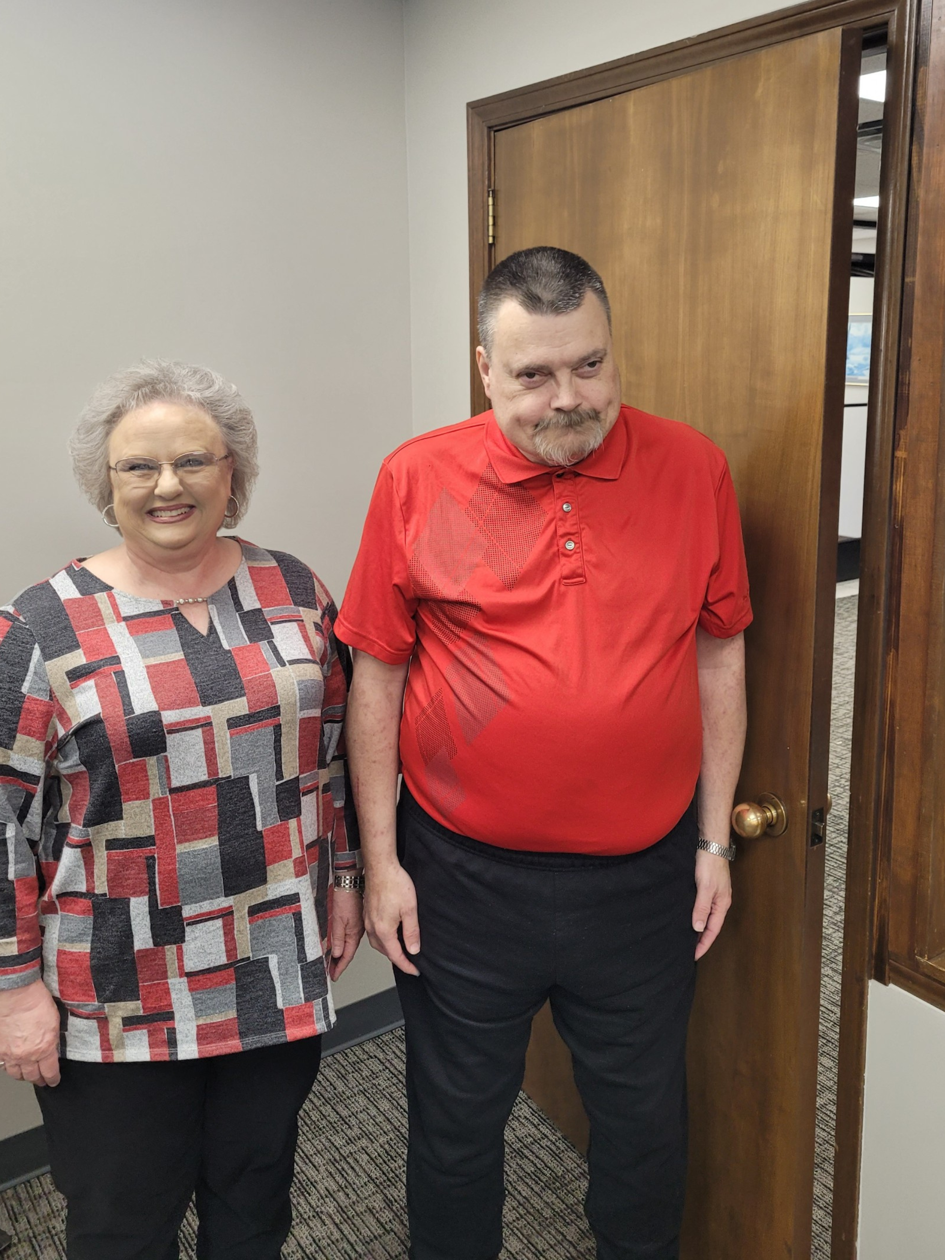 Older woman smiling with gray short hair wearing glasses and long sleeve blouse standing next to man with arms down by his side wearing a red short sleeve collared shirt